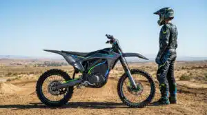 Motocross électrique : prix et modèles incontournables : Pilote en tenue devant une moto de motocross électrique futuriste gris et vert, sur une piste de terre sous ciel bleu.