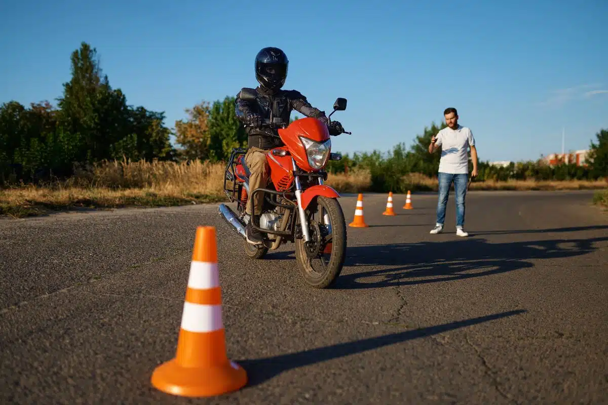 Élève motard en slalom entre les plots sur un plateau de moto-école, moniteur en arrière-plan chronomètre en main