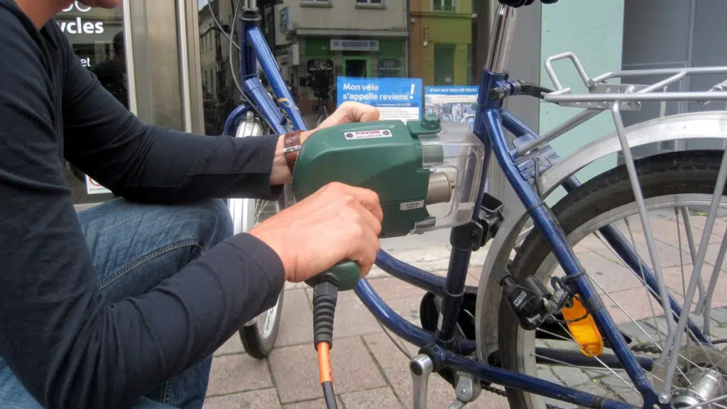 Opérateur agréé marquant un vélo au Bicycode avec une machine de gravure, affiche Mon vélo s'appelle reviens en arrière-plan